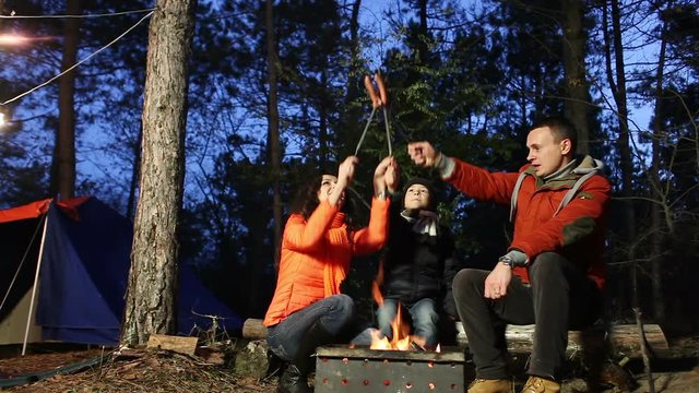 Happy family cooks sausages on campfire in the forest. Series.