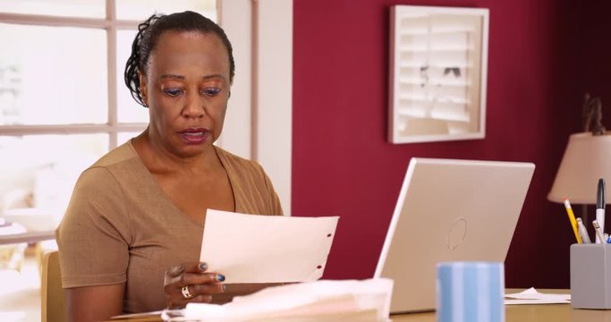 An older black woman uses her phone and laptop to do her taxes. An elderly African American woman uses her computer and mobile phone to do some accounting