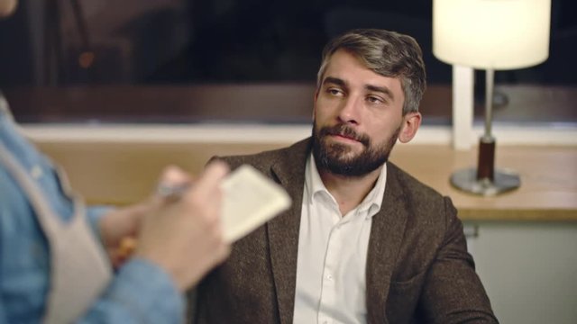 Elegant businessman sitting at cafe table and using digital tablet while waitress coming to take an order