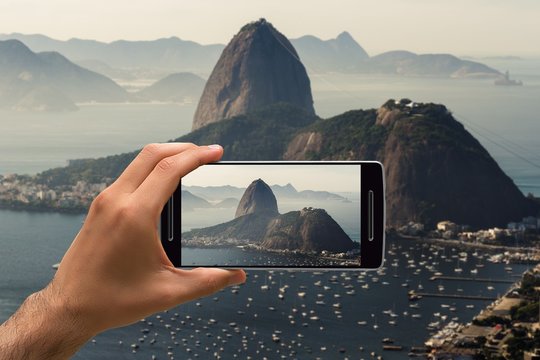 Man taking a photo with cellphone showing only the hand. It's a Sugarloaf Mountain in Rio de Janeiro, Brazil. It is a misty morning.