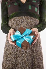 detail of brown gift box with blue ribbon in hands of woman with brown and green dress over white...