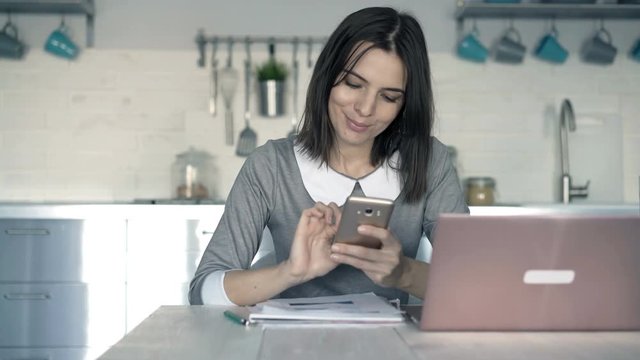 Young, happy businesswoman using smartphone sitting by table in office
