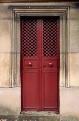 Red closed iron double door with door knobs and a hole pattern, surrounded by a stone wall. The door frame is rectangle and ornated with chiseled lines. The texture is made of small steel crosses.