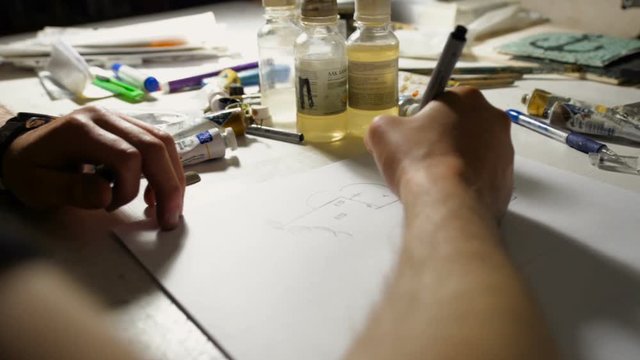 artist is painting by marker the sketch on art workplace with different painting tools, brushes, bottles, tubes under the lamp light