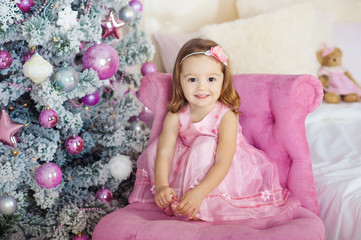 beautiful little girl sits and poses on a chair.   background  festive fir-tree   set of bright fires. Gently smiles. Xmas holiday concept