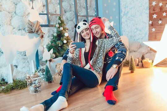 Beautiful young couple in funny hats sitting on the background of Christmas decorations and make selfie.