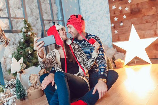 Beautiful young couple in funny hats sitting on the background of Christmas decorations and make selfie.