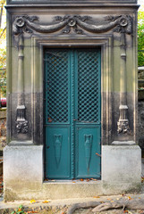 Rectangular turquise double door made of steel. Ornaments of vases, floral / cross patterns. The ornate chiseled stone door frame contains carved reliefs of torches. An old tomb / crypt at a graveyard