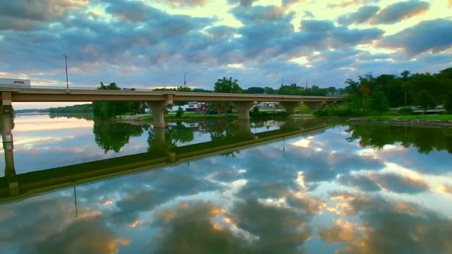 Flying under scenic bridge over tranquil waters reflecting moody sky at dawn.
