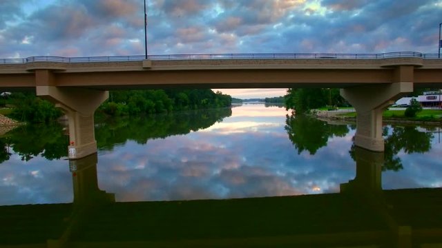 Flying under scenic bridge over tranquil waters reflecting moody sky at dawn.
