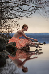 Ballerina in ballet pose sits above the lake on background of sk