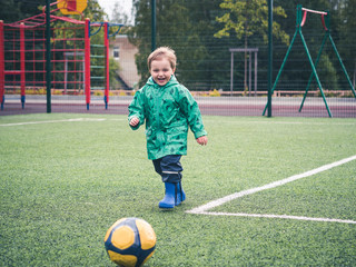 Cute little kid is playing with a yellow soccer ball on a children's on the playground. Sport, bad weather, rain.