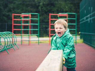 Fototapeta na wymiar Cute funny little kid playing on the playground in a beautiful green cloak with dinosaurs. Sport, bad weather, rain.