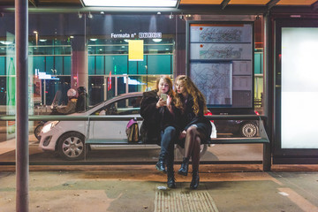 Two young beautiful caucasian women friends outdoor in the city night using smart phone hand hold sitting at bus stop - technology, social network communication concept - obrazy, fototapety, plakaty