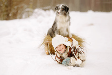 Cute little girl in a fur coat and boots smiling and pulling a sleigh on snow with hay on which the dog is sitting Outdoors