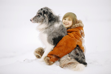 Smiling little girl child in the mother's fur coat hugging his friend the dog sitting in the snow in winter and smiling