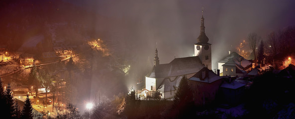 Winter in Slovakia. Old mining village. Historic church in Spania dolina. Fog at night. - obrazy, fototapety, plakaty