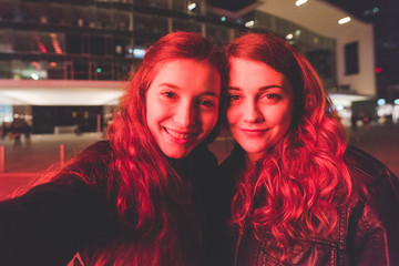 Fototapeta premium Two young beautiful caucasian women friends outdoor in the city night having fun taking selfie illuminated by red light -social network, sharing, vanity concept
