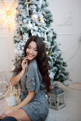 Beautiful smiling girl in a gray dress sits near a Christmas tre