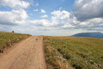 Fototapeta na wymiar The simple landscape with the earth-road, the distant mountain, the field with green grass and the sky with clouds. This photo was taken in Crimean Mountains, on South Demerdzhi mountain.