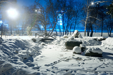 Snowy city park in light of lanterns at evening.Snow-covered trees and benches,footpath in a fabulous winter night park.Winter landscape