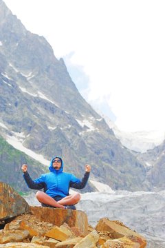 Young man in sunglasses and a hooded jacket on the edge of a cliff doing yoga, sitting in Lotus pose with raised hands, against the background of snow-capped peaks. Sport and active life concept