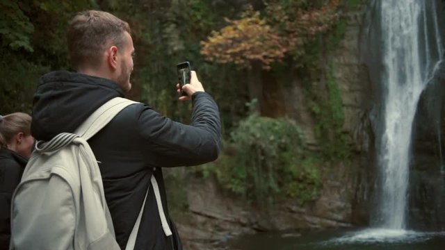 Young man taking a photo of a beautiful waterfall with a smartphone slow motion
