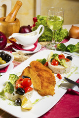 Portion, two pieces of deep fried fish rolled in bread crumbs and corn flour; side dish of boiled rice and broccoli; fresh cherry tomatoes, olives and lemon beside a glass of wine and sauce.