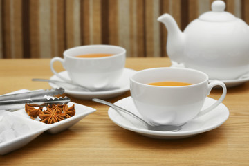Two cups of tea, tongs and plate with sugar and spices on wooden table
