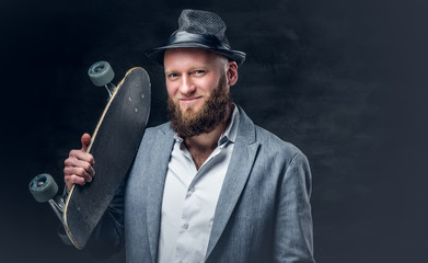 Bearded male in a suit and felt hat holds skateboard.