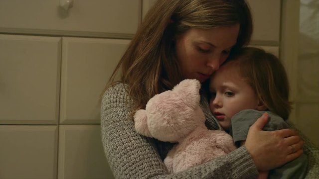 Sad, scared young girl with a stuffed animal being held by her mother in her room