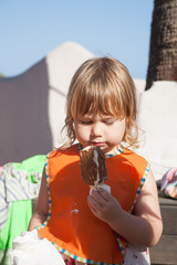 portrait of three years old blonde happy child with orange bib eating chocolate and white popsicle...