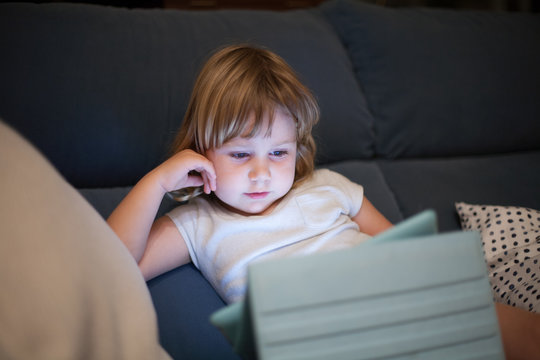 blonde three years old baby shirt and shorts, sitting comfortably in sofa inside home at night reading and watching digital tablet, face illuminated by the light of the screen
