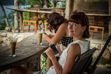 Fototapeta na wymiar Best friends having fun, laughing. Tropical cafe on the cliff in Nusa Lembongan, Indonesia. Girls drinking ice coffee.