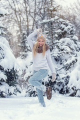 Attractive young blonde girl walking in winter forest. Pretty woman in wintertime outdoor. Wearing winter clothes. Knitted sweater, scarf, hat and mittens.