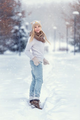 Fototapeta na wymiar Attractive young blonde girl walking in winter forest. Pretty woman in wintertime outdoor. Wearing winter clothes. Knitted sweater, scarf, hat and mittens.