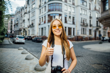 Fototapeta na wymiar Outdoor summer smiling lifestyle portrait of pretty young woman having fun in the city in Europe in evening with camera travel photo of photographer Making pictures in hipster style sunglasses