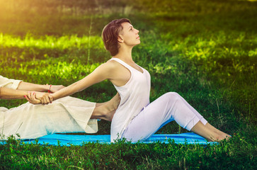 Young man and woman dressed in a white robe doing thai massage with yoga exercises sitting on the mat. Sunny summer park with green lawn in the background