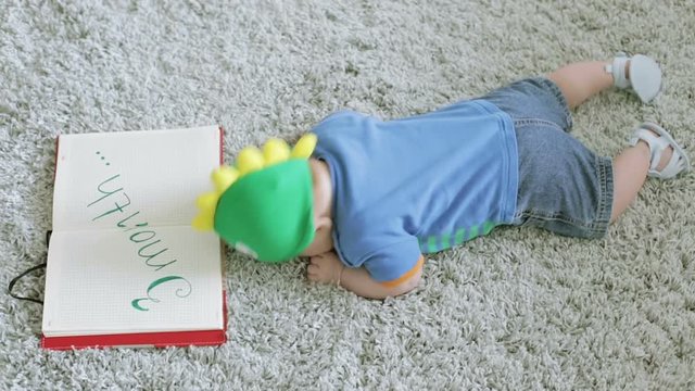 Three-month baby in a green dragon form hat looks in a notebook lying on the floor on his stomach.