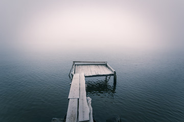 Frozen wooden pier on a cold foggy river and smooth reflection on water. .