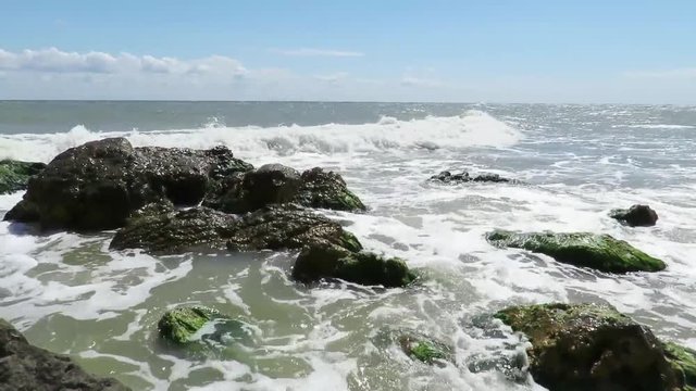 Sunny summer day at sea with scarce clouds at the horizon and  waves hitting shoreline rocks creating foam , in slow motion