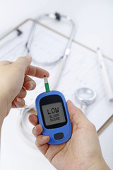 Hand holding a blood glucose meter measuring blood sugar, the background is a stethoscope and chart file