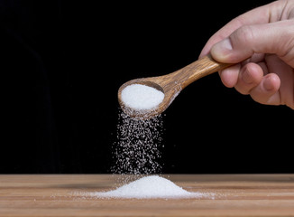 Person is pouring sugar or salt with a wooden spoon. A pile of ingredient is on the wooden surface.