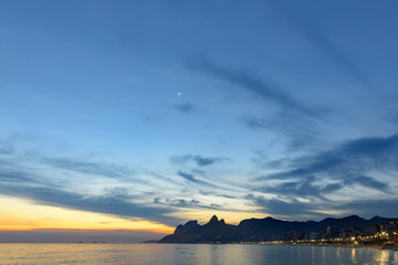 Arpoador, Ipanema and Leblon beaches in Rio de Janeiro during dusk with its lights, moon, and sky and the hill Two brothers and Gávea stone in the background