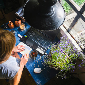 Mid adult woman sitting at desk, with laptop, looking at notebook, overhead view