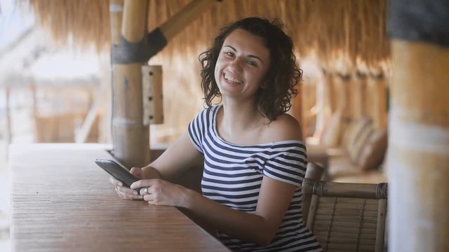 Tanned young girl sitting at the outdoor bar under a straw umbrella holding a tablet computer in the hands and fervently laughs
