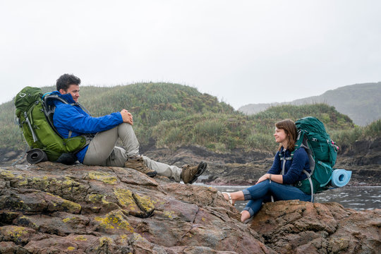 Young couple with backpack, sitting on rocks, Constant Bay, Charleston, South Island, New Zealand