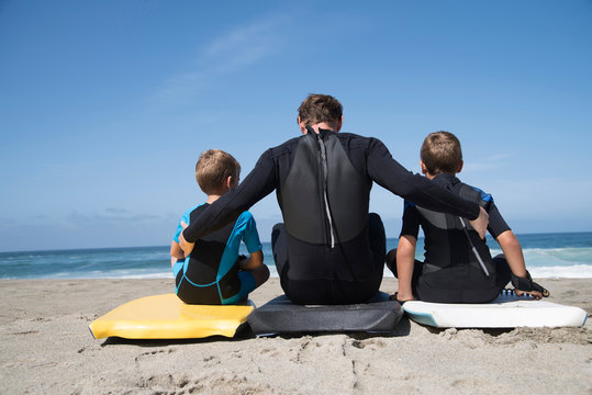 Rear view of man and two sons sitting on bodyboards, Laguna Beach, California, USA