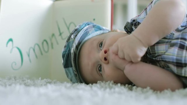 Three-month baby in the checkered cap lying on the floor on a gray carpet and sucks his arms.