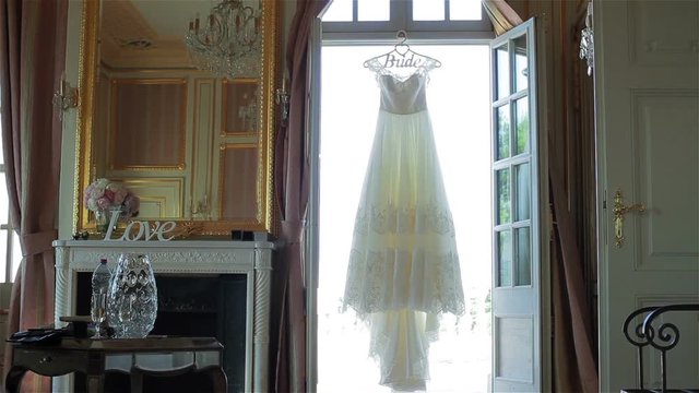 Wedding dress hanging in doorway light waiting for bride tracking pan. Lace decorated white bridal gown on hanger in window luxury room interior decor golden elements. Morning of bride preparations